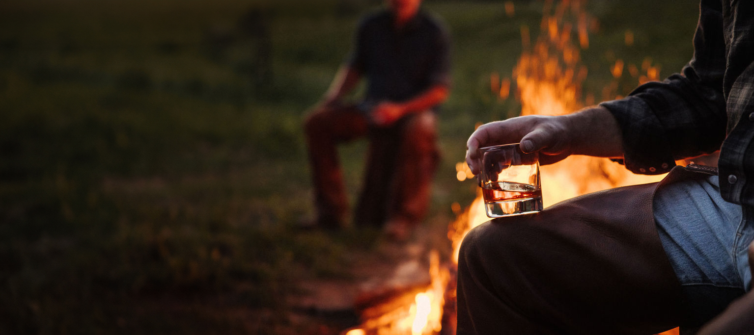 A man holding a glass of Stetson Bourbon by a campfire